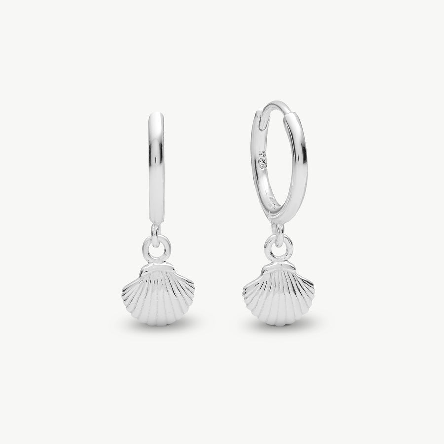 A pair of sterling silver huggie hoop earrings with dangling scallop shell charms, shown on a light gray background.