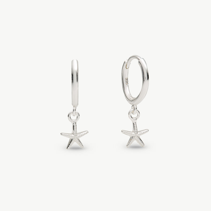 A pair of silver huggie hoop earrings with dangling starfish charms, shown on a clean off-white background.