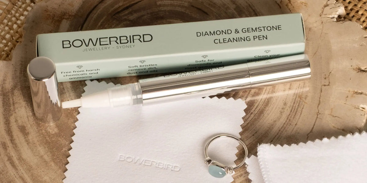 Bowerbird Diamond & Gemstone Cleaning Pen set with its mint green box, silver pen, white microfiber cloth, and a sample ring on a rustic wood slice background.