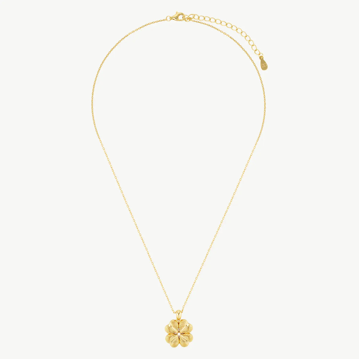 A ridged four-leaf clover charm with a cubic zirconia centred gemstone hanging from a fine yellow gold cable chain on a cream background 