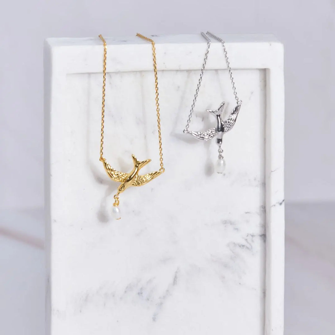 Two bluebird necklaces on a marble surface, one gold and one silver.