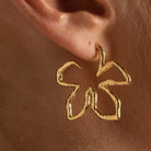 A close-up of a person's ear wearing a single textured gold hoop earring shaped like a stylized flower or abstract loop.