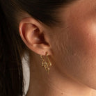 A close-up of a model's ear wearing a delicate gold hoop earring with three dangling looped leaf-like charms.
