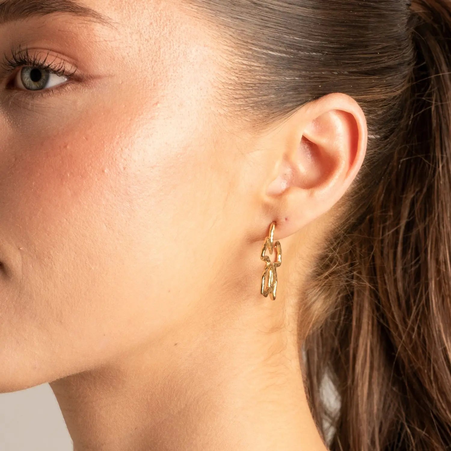 A close-up of a woman's ear wearing a delicate gold earring with two interlocking curved hoop-like links.