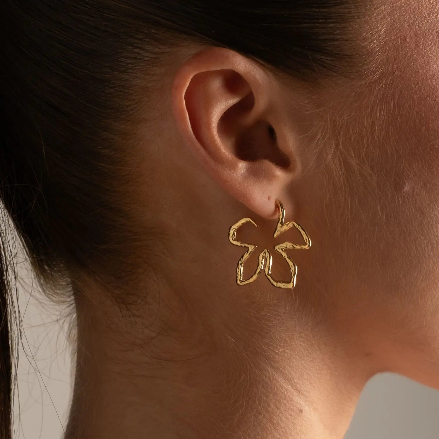 A close-up of a model's ear wearing a delicate gold hoop earring with a small, openwork butterfly or flower-like motif.