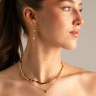 A close-up of a woman wearing layered gold necklaces and a dangling gold earring with a pearl accent, shown against a plain light gray background.