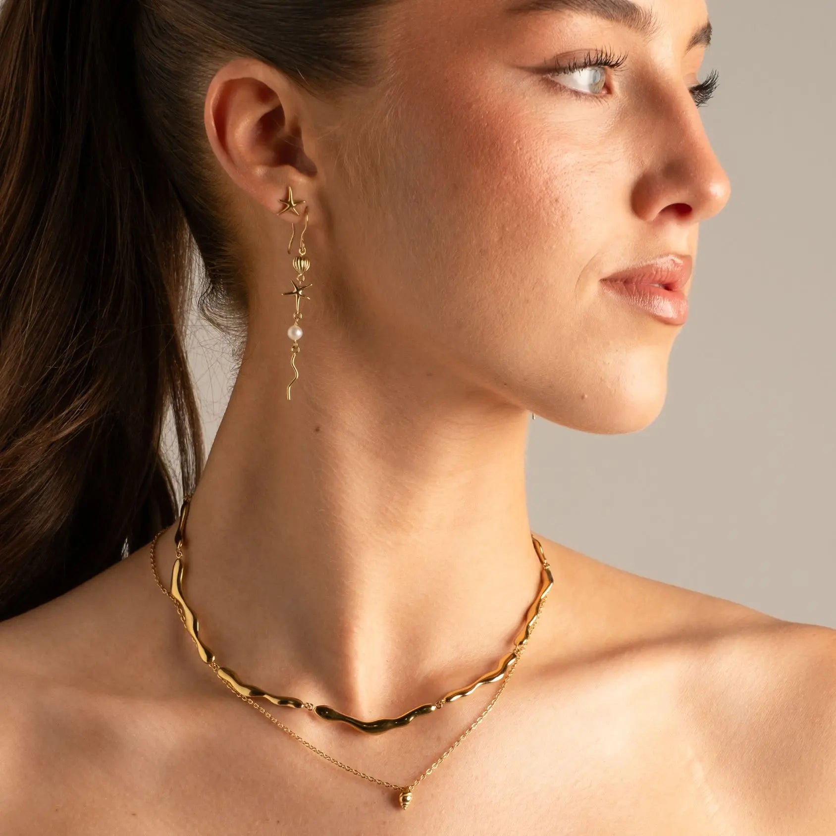 A close-up of a woman wearing layered gold necklaces and a dangling gold earring with a pearl accent, shown against a plain light gray background.