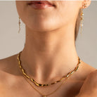 Close-up of a model wearing a gold wavy chain necklace layered with a delicate gold chain necklace, along with matching dangling earrings.