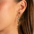 A close-up portrait of a woman wearing a delicate gold bracelet with green floral accents on her wrist, along with matching gold earrings and rings, set against a solid neutral beige background.