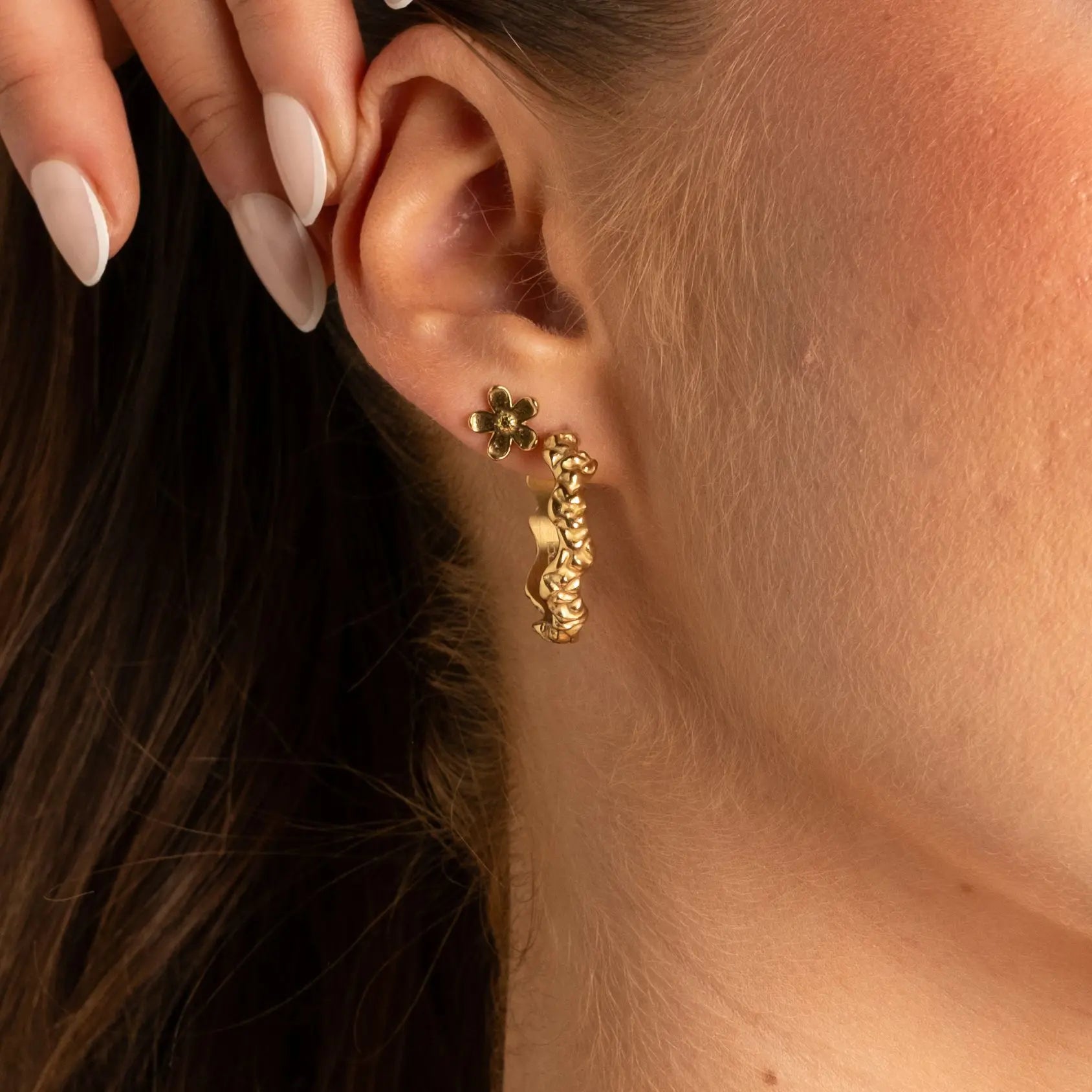 A close-up of a model's ear wearing a gold huggie hoop earring with a dangling textured gold charm and a small green flower stud.