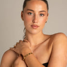 A close-up portrait of a woman wearing a delicate gold bracelet with green floral accents on her wrist, along with matching gold rings and earrings, set against a solid neutral beige background.