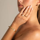 A close-up lifestyle shot of a woman wearing a delicate gold flower bracelet on her wrist, gold rings on her fingers, and a small gold earring, set against a soft beige background.