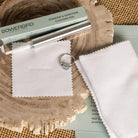 Bowerbird diamond and gemstone cleaning pen kit with a silver metal pen, a light green box, two white microfiber cloths, and a small ring with a blue stone, arranged on a rustic wood slice and burlap background.