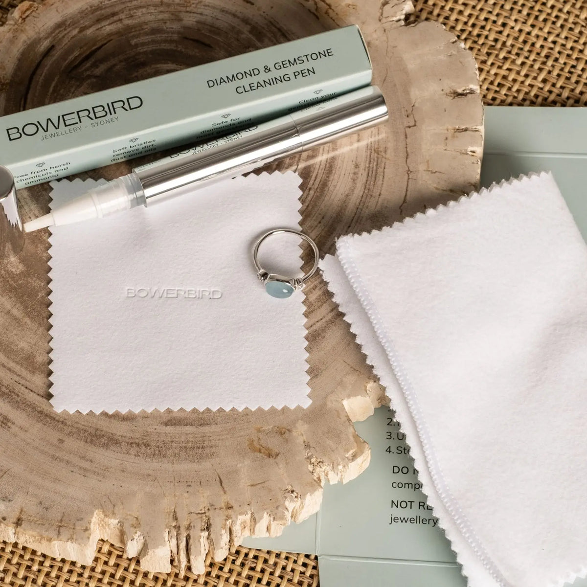 Bowerbird diamond and gemstone cleaning pen kit with a silver metal pen, a light green box, two white microfiber cloths, and a small ring with a blue stone, arranged on a rustic wood slice and burlap background.