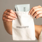 A close-up of a person holding a white Bowerbird jewelry pouch with the brand name printed in black. The person's hands with manicured nails are visible holding the pouch open, which reveals a light blue jewelry box inside.