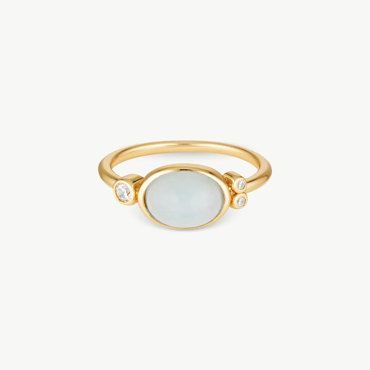 A gold ring featuring a large oval pale blue-gray stone in the center, flanked by two small clear gemstones on the band, set against a plain off-white background.