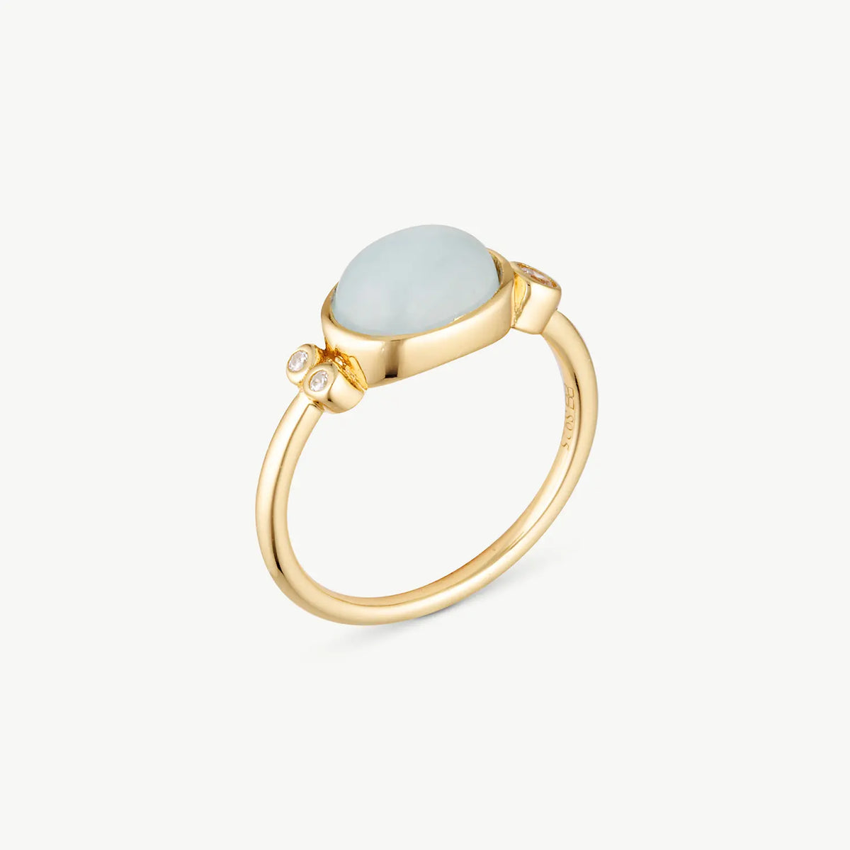 A gold ring featuring a smooth, oval-shaped light blue stone and a small round clear accent stone on the band, set against a clean off-white background.