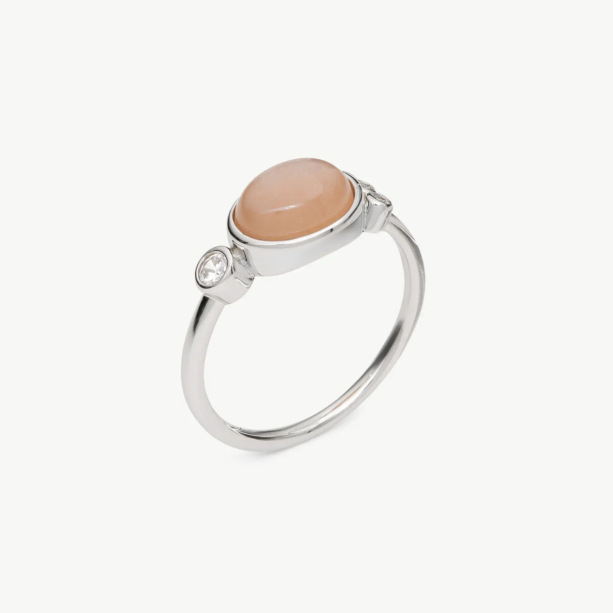 A silver ring featuring a smooth, oval peach-colored cabochon stone and a small clear accent gemstone on the band, set against a plain off-white background.