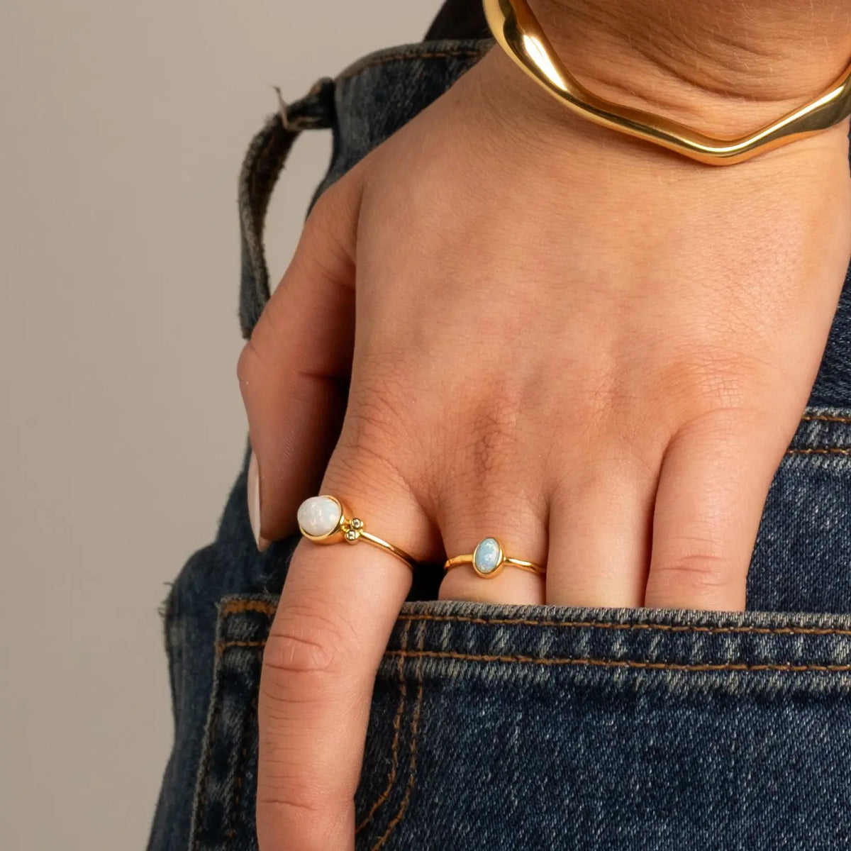 Picture of hands in a denim jean pocket with two rings on the fingers and a gold cuff bracelet 