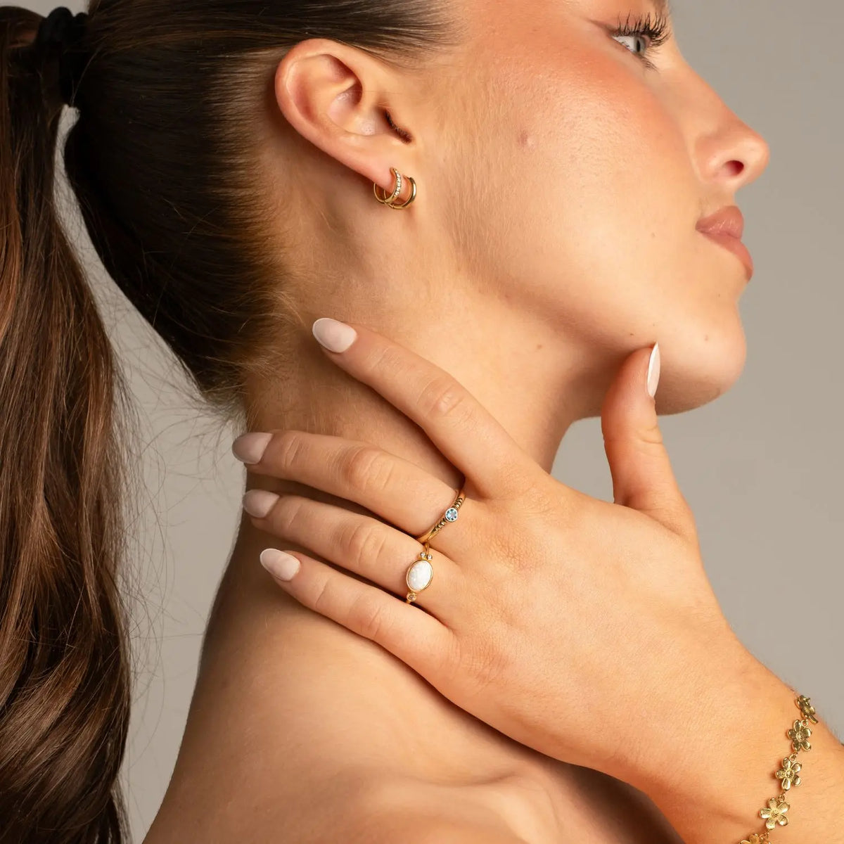 Side profile of a woman holding her neck with two earrings in her ears. She's looking off into the distance. She has two rings on her fingers featuring a blue and a white stone, and a flower bracelet around her wrist. 