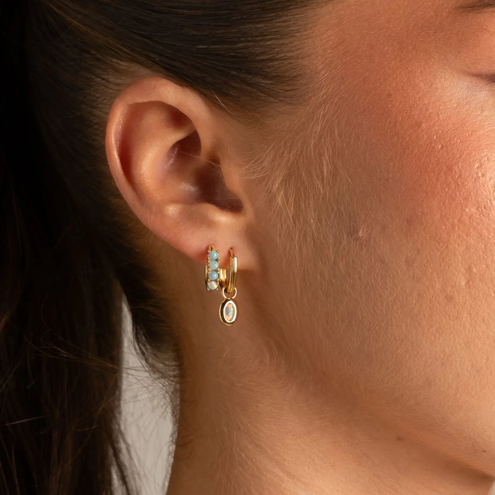 A close-up of a person's ear wearing a pair of gold hoop earrings with small light blue and white accents and a dangling oval charm.
