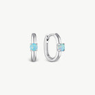 A pair of silver huggie hoop earrings, one shown from the front and the other from the side, each featuring a round-cut light blue opal gemstone in a four-prong setting, on a clean off-white background.