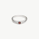 A silver ring with a textured beaded band and a small round red gemstone in the center, shown on a plain light gray background.
