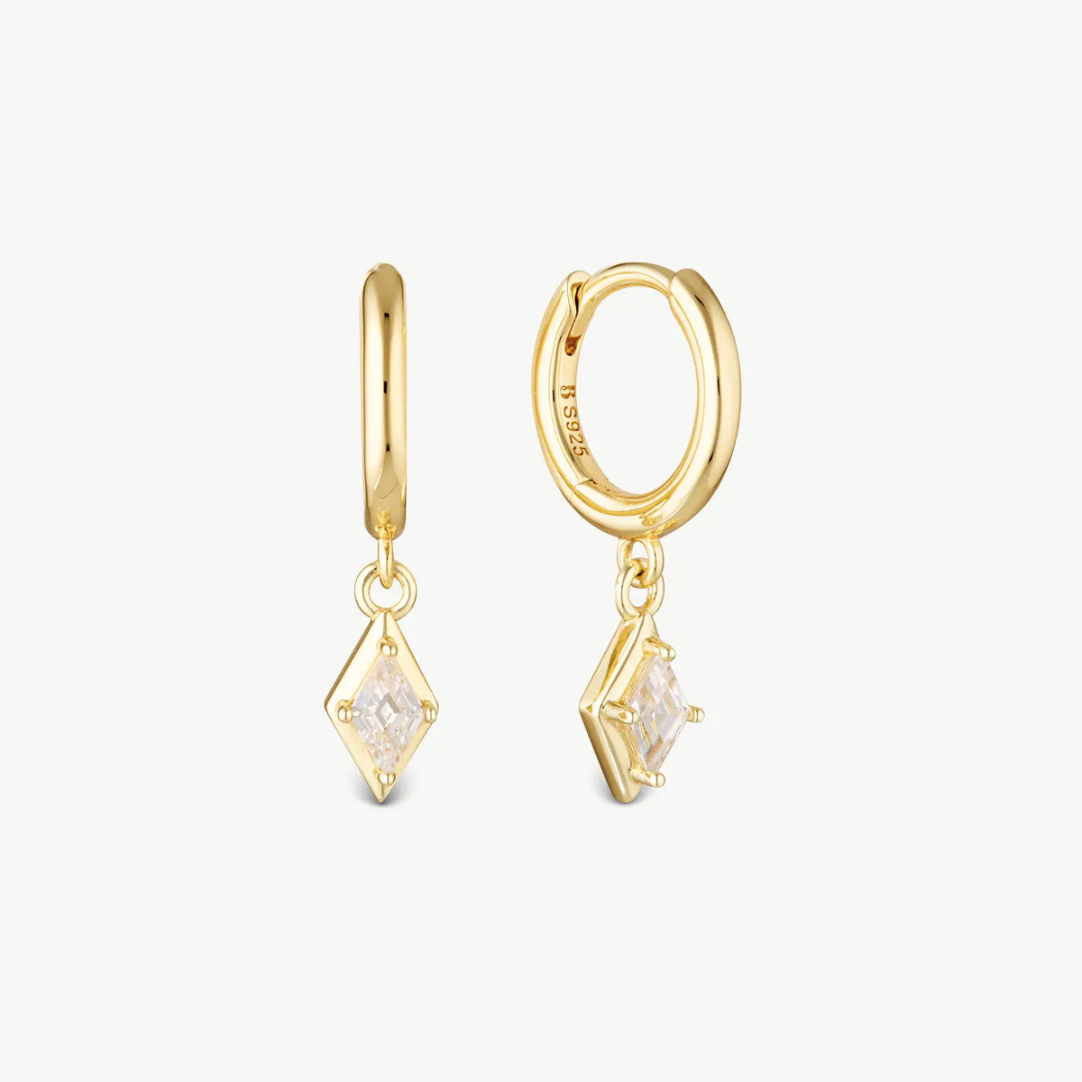 A pair of gold huggie hoop earrings with dangling diamond-shaped clear gemstone charms, shown on a light off-white background.
