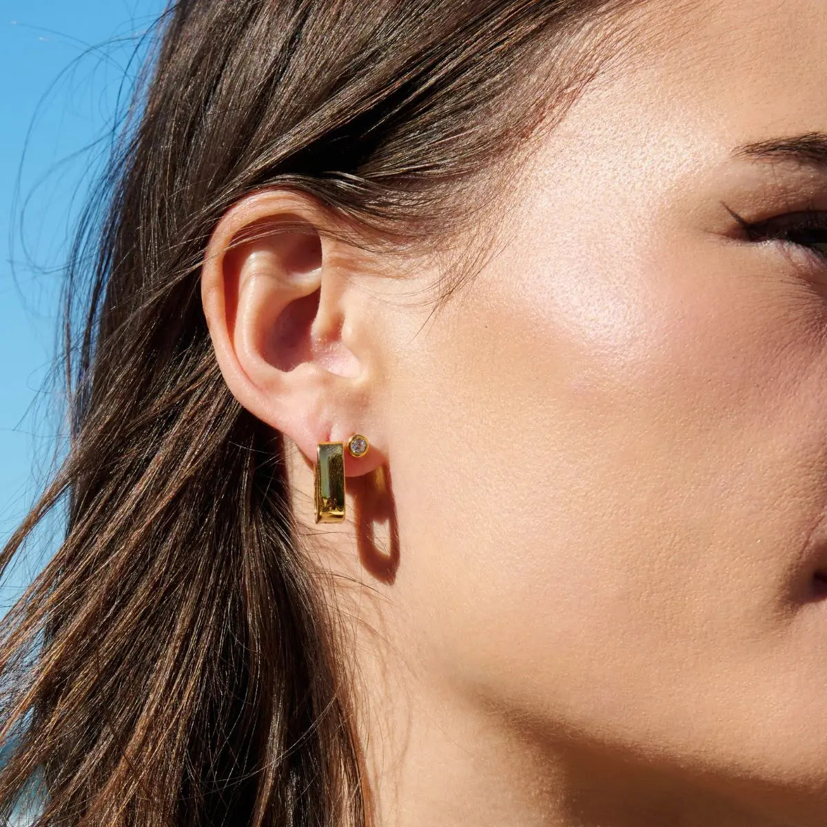 Close-up of a woman's ear wearing a gold earring with a blue sky background