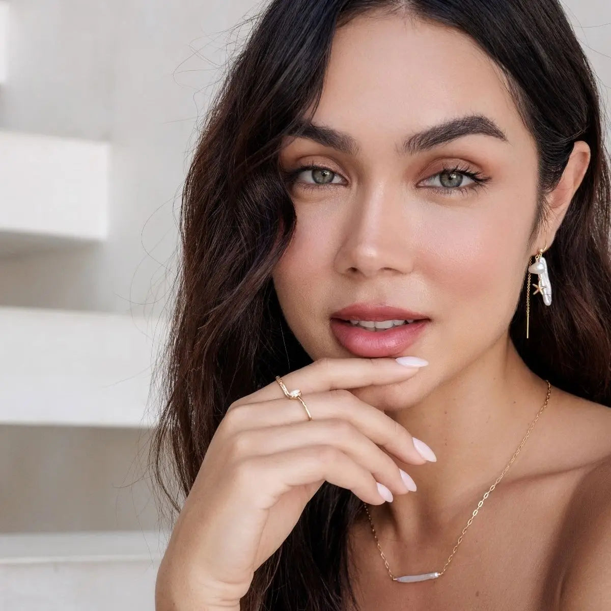 Woman with a neutral expression, wearing earrings and a necklace, with a blurred background.