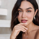 Woman with a neutral expression, wearing earrings and a necklace, with a blurred background.