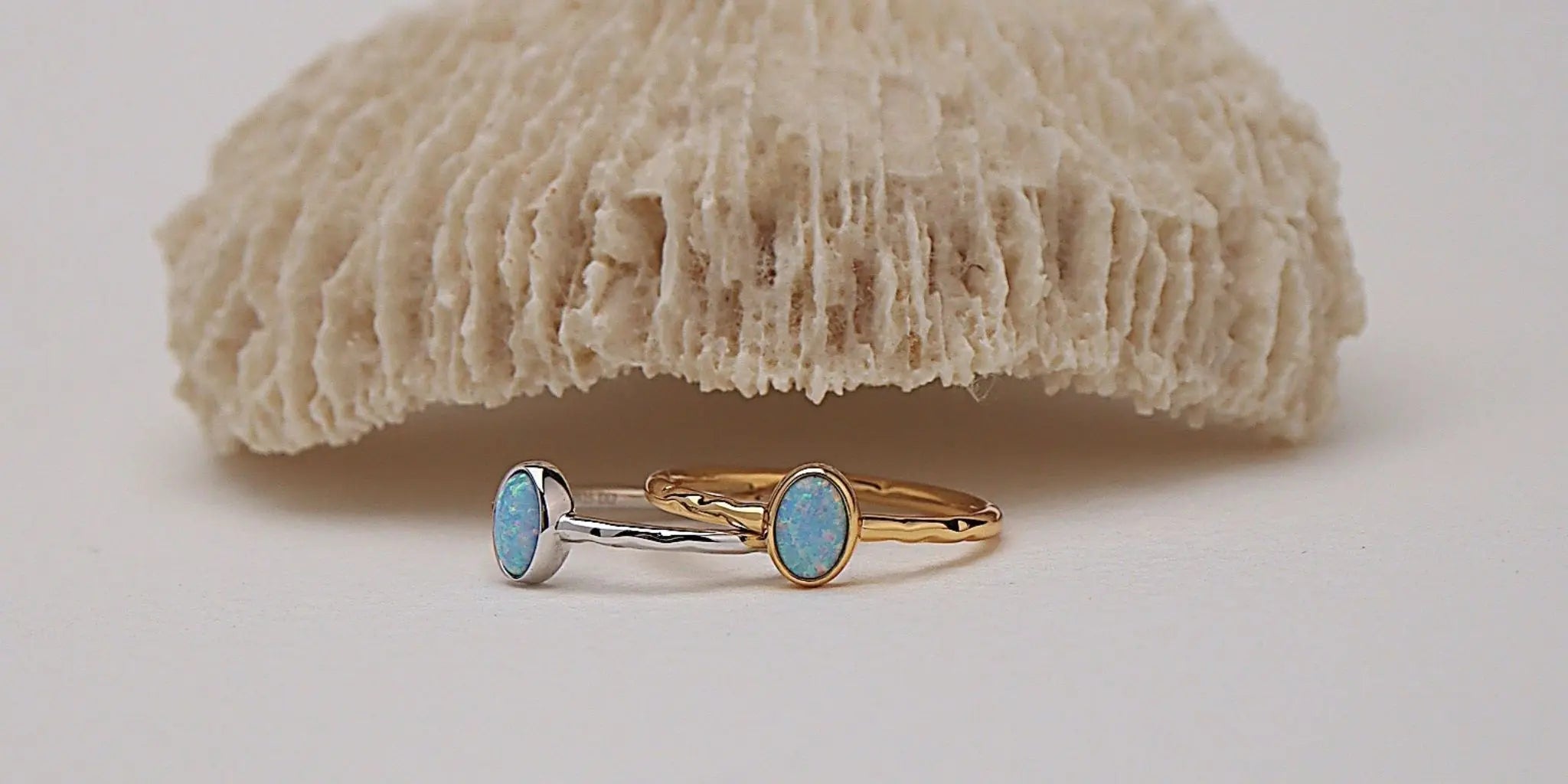 Two slim rings with oval light blue stones, one in silver and one in yellow gold, displayed in front of a textured beige coral-like prop on a white background.