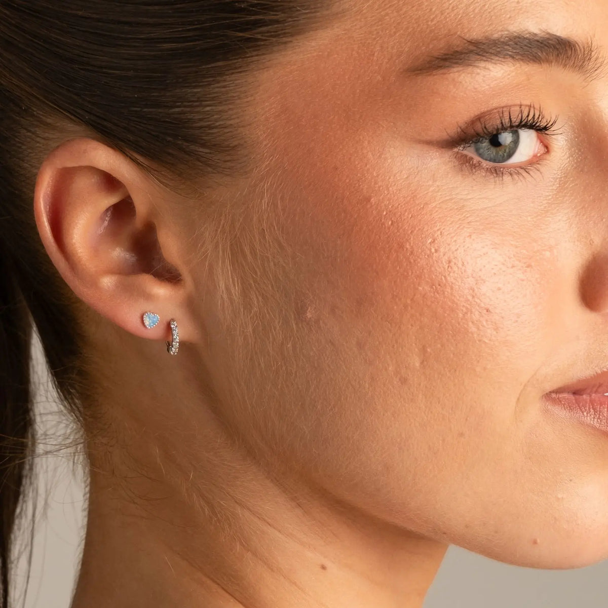 Close-up of a woman's ear wearing a small silver hoop earring with a light blue heart-shaped crystal inset.