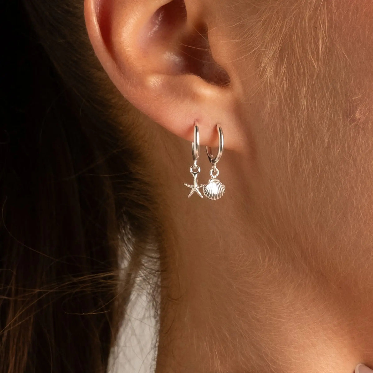 A close-up of a person's ear wearing a pair of silver hoop earrings with dangling starfish and shell charms.